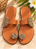 A pair of tan leather slide sandals with colorful beaded toe loops and matching beaded arch straps, displayed on a textured stone surface with tropical leaves and a flower.