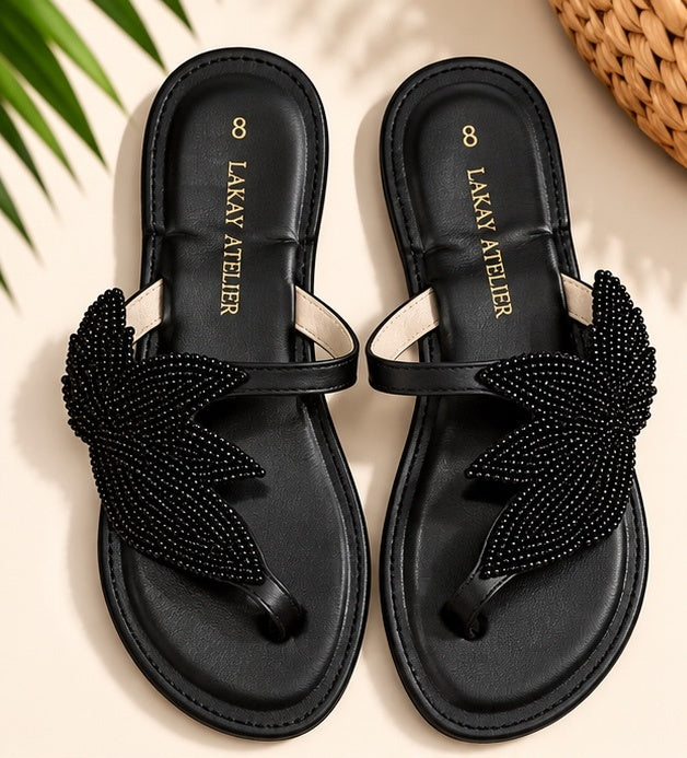 A pair of black flat thong sandals with beaded leaf-shaped straps, displayed on a light beige background with decorative green leaves and a woven basket corner.