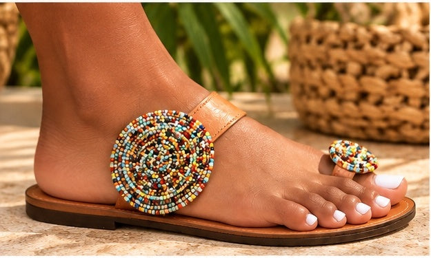 A close-up of a person wearing tan leather slide sandals with colorful beaded oval and round embellishments on the straps, shown outdoors on a stone surface with green foliage in the background.