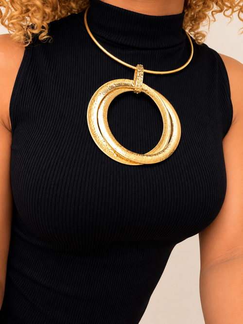 A close-up of a model wearing a chunky gold statement necklace with a large textured double-ring pendant over a black ribbed turtleneck top.