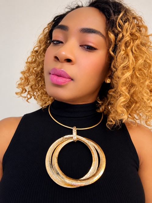 A close-up of a woman with curly blonde hair wearing a large textured gold double-ring pendant necklace over a black turtleneck top.