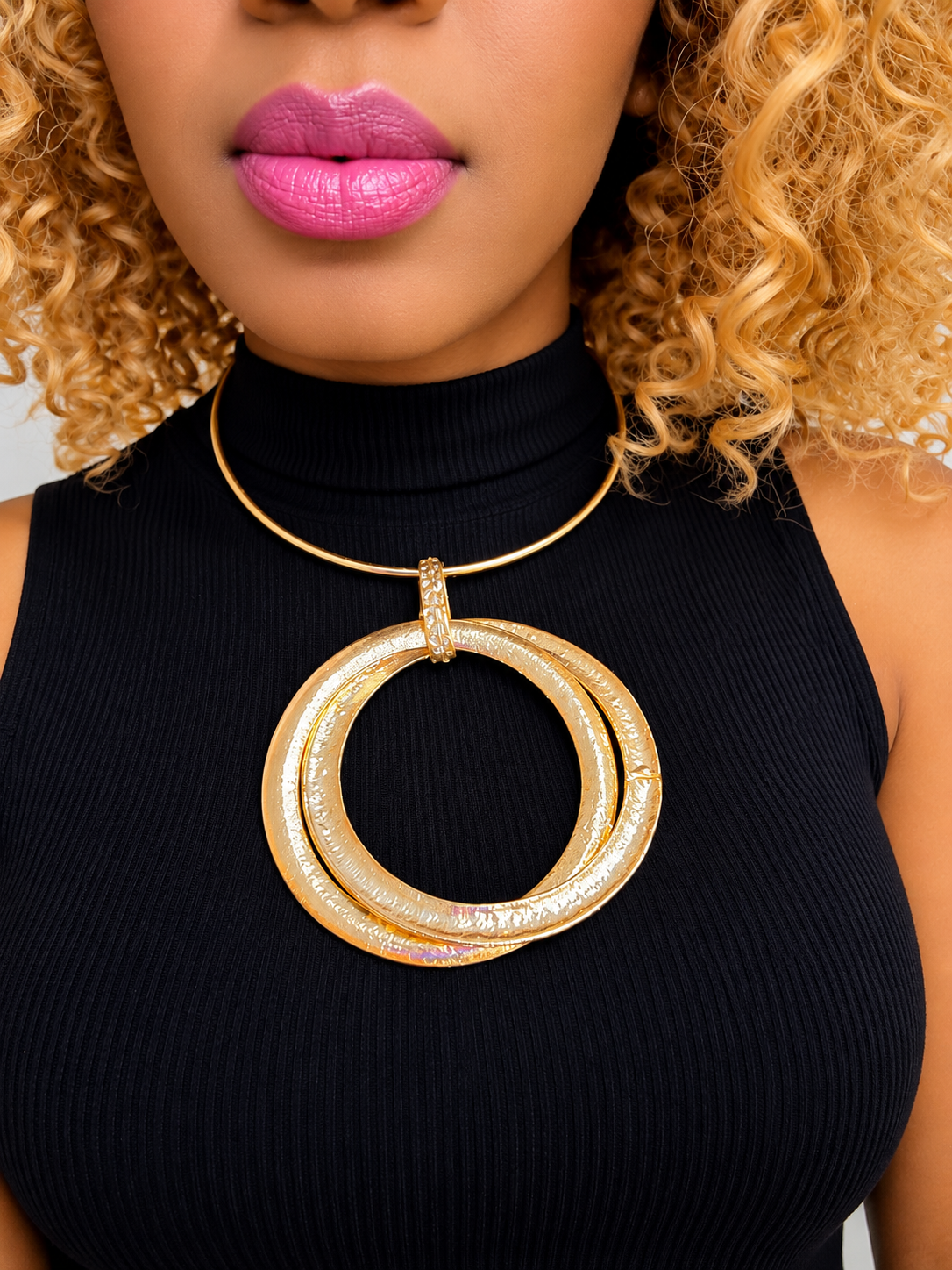 A close-up of a model wearing a chunky gold statement necklace with two interlocking textured rings and a small rhinestone-encrusted top loop, paired with a black ribbed turtleneck top.